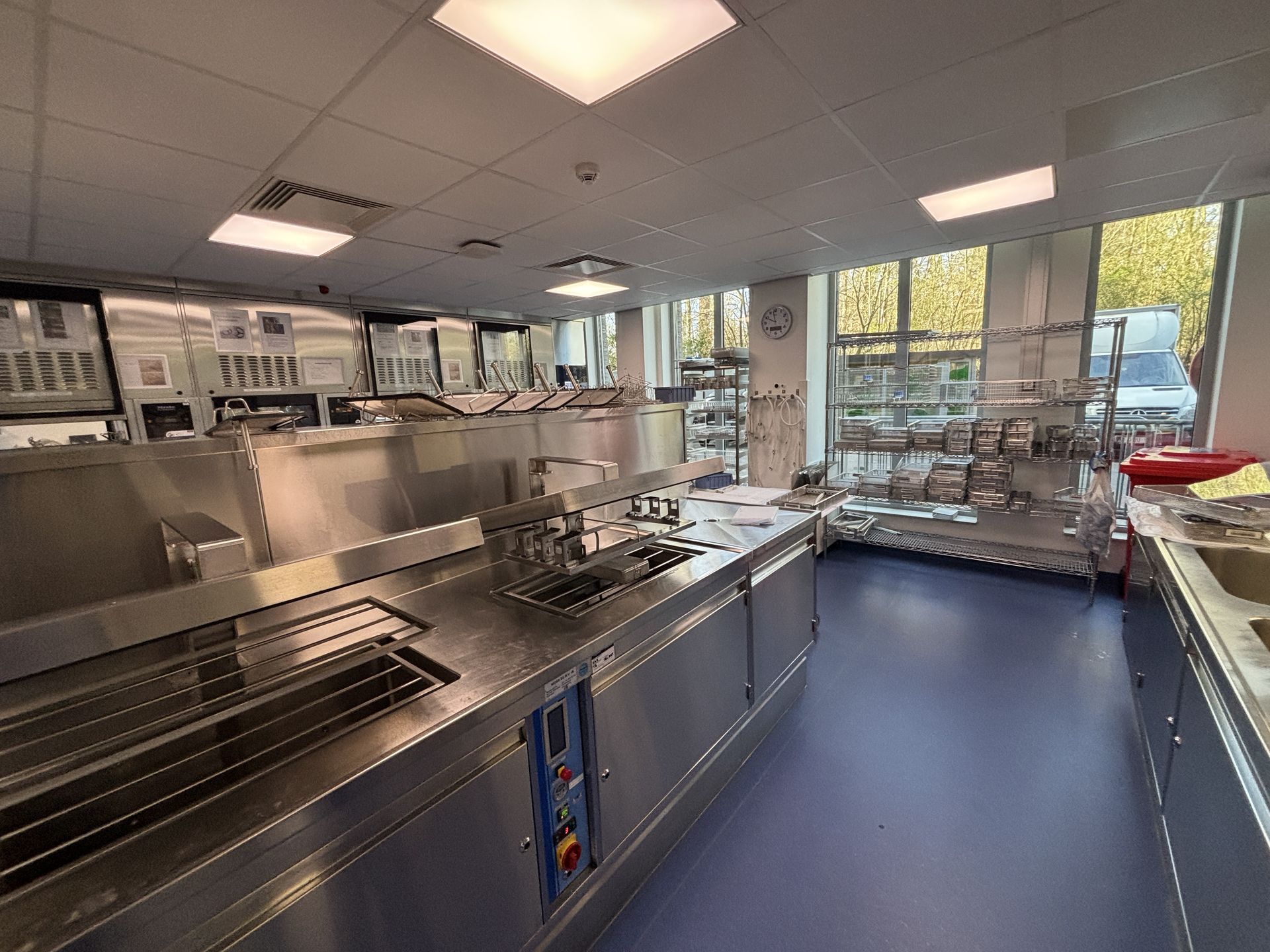 A wide, clean hospital CSSD kitchen featuring stainless steel appliances and counters. The room has a bright blue floor and white walls, with large square light panels on the ceiling. A long prep island with sinks and stovetops runs down the center, . Windows look out onto a parking area on the right.