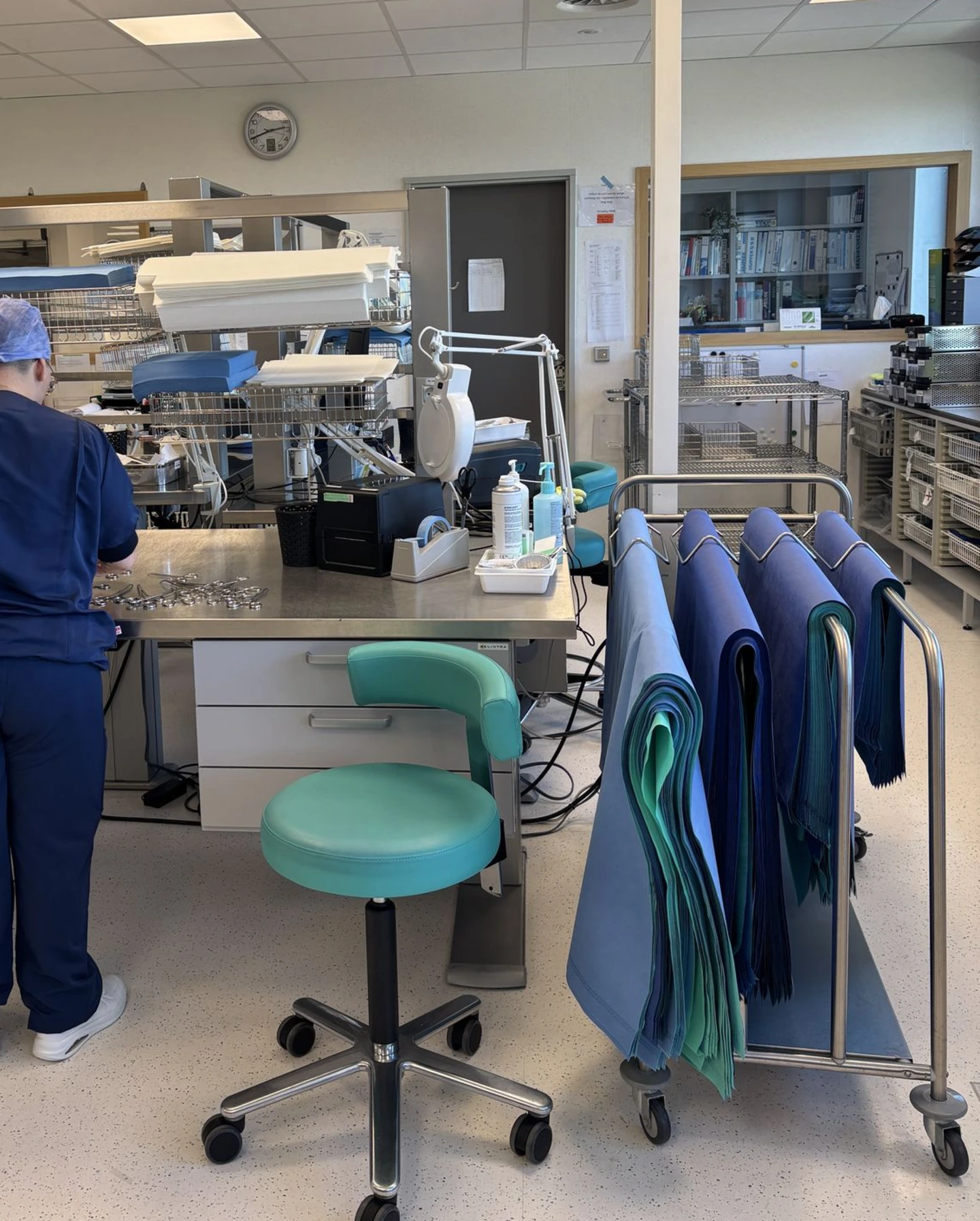 CSSD-wrapping workstation Medical professionals in blue scrubs and surgical caps are working at specialized, well-lit stations, processing and inspecting medical instruments. A trolley with folded blue and green sterile drapes is visible, highlighting the preparation process for surgical procedures.