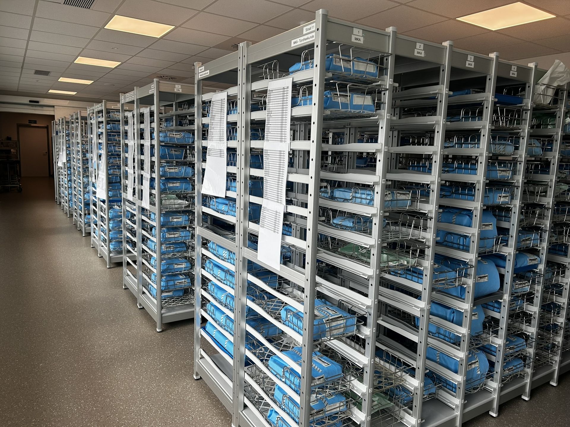A well-organized hospital sterile supply room showing rows of metal shelving units. The shelves are filled with blue-wrapped, sterilized surgical instrument trays,